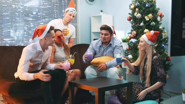 A group of seven young adults wearing Santa hats and party masks enjoy colorful drinks while seated around a coffee table near a decorated Christmas tree in a modern lounge.