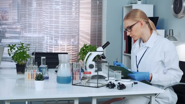 A female scientist wearing a lab coat and gloves is writing notes on a clipboard while seated at a laboratory table with scientific equipment and plants indoors.
