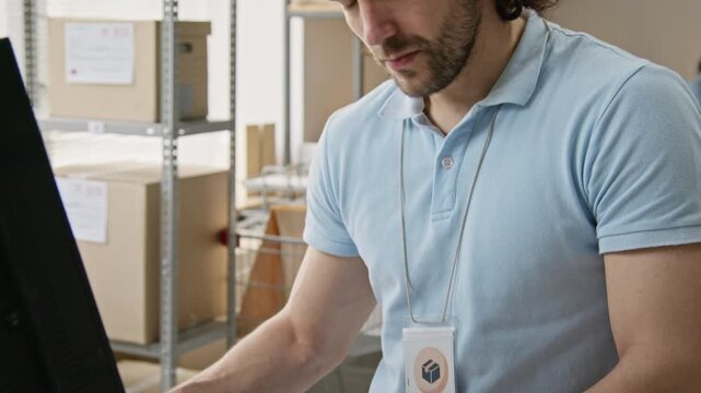 Caucasian young order collector typing on computer keyboard in warehouse. Ideal for e-commerce software marketing, logistics management, inventory tracking, delivery service promos