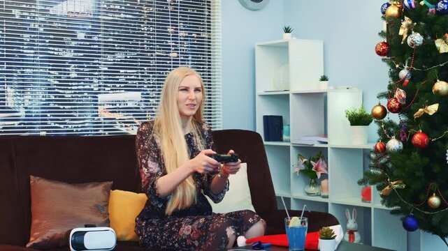 A blonde woman in a patterned dress is smiling and playing with a gaming controller while seated on a sofa in a modern living room decorated for Christmas with a tree and city view.