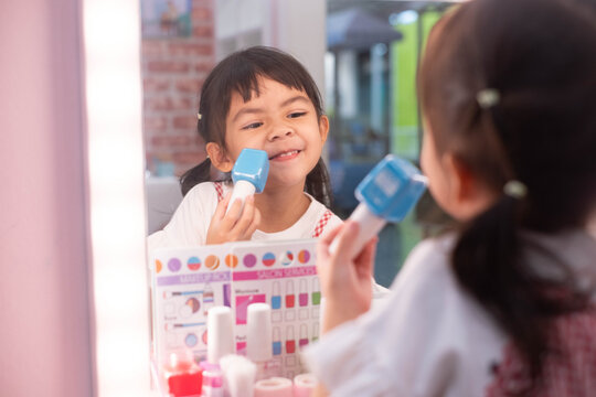 Happy young girl pretending to apply toy makeup in front of a mirror, smiling during creative beauty roleplay that encourages imagination, confidence, self-expression, learning, and childhood fun.