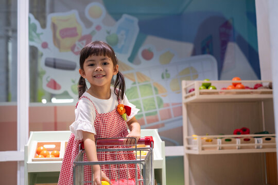 Young girl pushing a shopping cart in a pretend grocery store, choosing toy vegetables while learning about healthy food, nutrition, independence, creativity, and decision making through play.