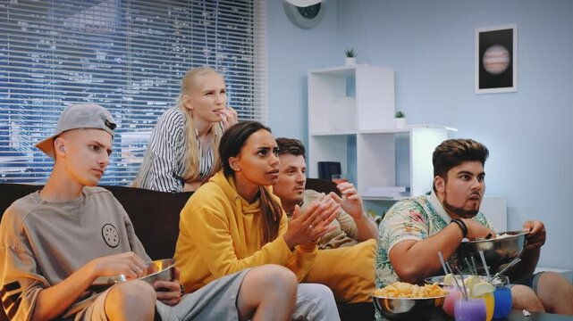 A group of eight young adults sit on a sofa in a modern living room, sharing a moment of disappointment while watching something off-screen during daytime.
