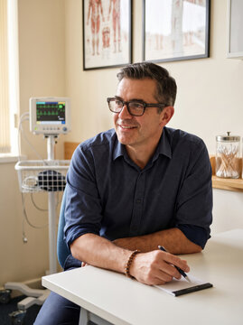 Friendly male doctor sitting at his desk in a modern consultation room, illustrating approachable primary healthcare and patient trust.
