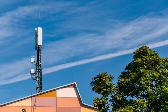 Cellular 5G mobile network antenna mast on roof of colorful modern building next to green trees against clear blue sky, telecommunication and digital infrastructure concept