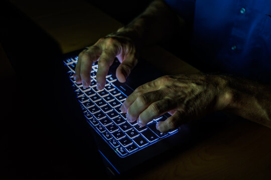 Close up of hands typing on illuminated laptop keyboard in dark room, cyber crime hacking and IT security concept