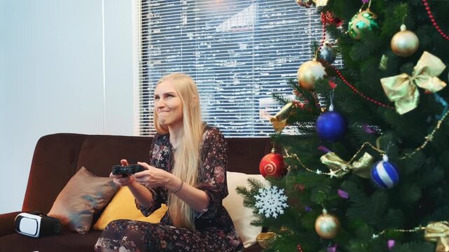 A blonde woman wearing a dark patterned dress celebrates winning a video game while seated on a sofa next to a decorated Christmas tree in a living room with city view.