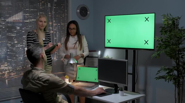Three designers collaborate on a 3D model using multiple monitors and laptops in a nighttime office studio with city skyline views and modern decor.