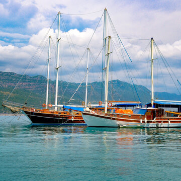Traditional wooden gulet yachts anchored on calm turquoise sea against scenic coastal mountains, square orientation shot featuring sky and water copy space for vacation concepts.