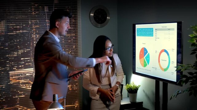 Three business professionals discuss data and infographics on a large digital screen in a modern office with cityscape views during daytime.