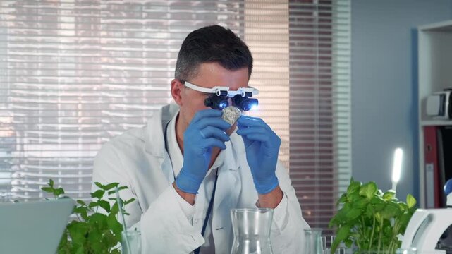 A middle-aged scientist wearing a lab coat and blue gloves examines a light-colored stone sample using magnifying glasses in a well-lit modern laboratory.