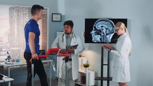 A male athlete on a treadmill is assessed by two doctors reviewing his brain MRI scan in a modern sports laboratory setting during daytime.