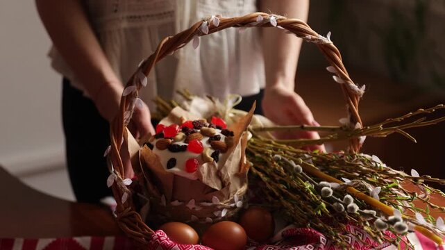A woman adjusts the paska bread inside a decorated Easter basket filled with eggs and willow branches on a wooden table.