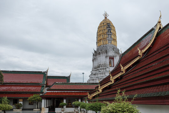 Low-angle exterior view of the historic golden prang stupa tower towering above the multi-tiered glazed ceramic tile roofs of the courtyard cloister at Phuttha Chinnarat temple in Phitsanulok.