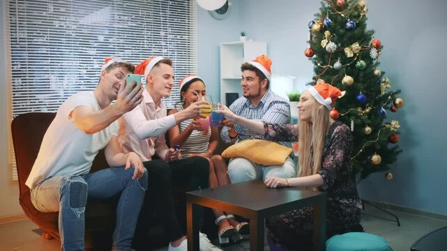 Seven young adults wearing Santa hats socialize indoors around a coffee table with drinks and smartphones by a decorated Christmas tree.
