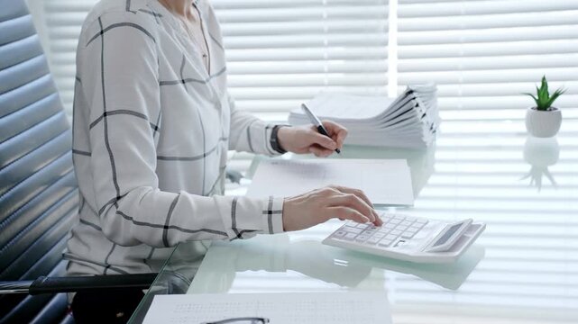 Professional accountant's hands working on financial calculations using a calculator and writing notes on documents, managing business tasks and verifying data in an office environment