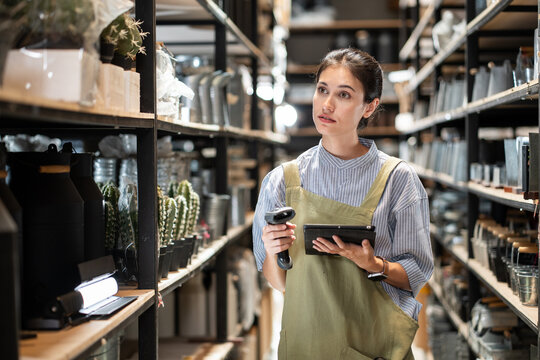 A worker uses a digital tablet and barcode scanner to check inventory while walking between shelves filled with goods in a warehouse, conveying focus and diligence.