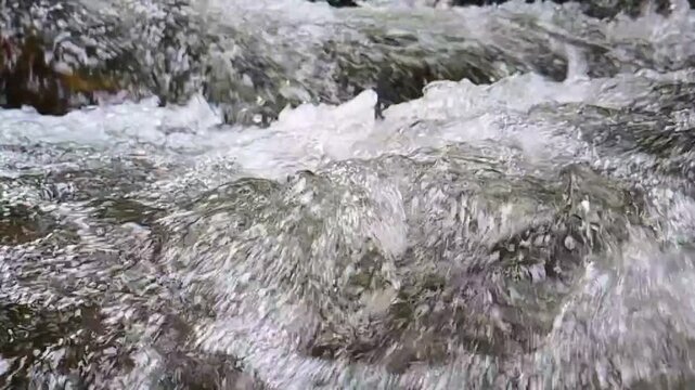 HD slow-motion close-up video of powerful water rushing between natural rocks and splashing over the rocky stream bed. The camera captures detailed droplets