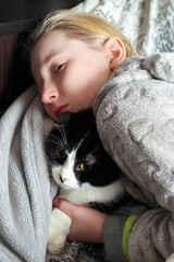 Young blonde girl cuddling black and white cat under soft grey blanket © parovysh