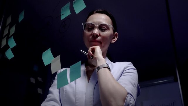Woman wearing clear frame glasses standing in front of a glass wall covered with sticky notes and drawing a mind map, concentrating on planning a business strategy