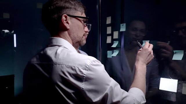 Business professionals brainstorming new ideas and strategy, collaborating on project development using sticky notes on a glass board in a dark modern office meeting room