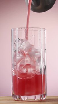 Watermelon juice fills rotating tall clear glass with ice on pink background for summer mocktail.