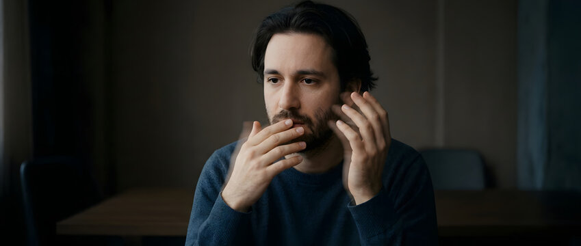 Concerned adult man, thinking deeply with hands near mouth, dark blue sweater and facial hair, dimly lit indoor home office interior, perfect for mental health awareness marketing and insurance.