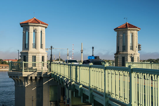 Two Mediterranean-style towers rise above the Bridge of Lions in Saint Augustine, Florida. Cars cross the green drawbridge beside blue water under a clear sky.