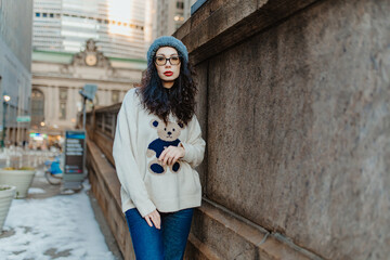 Woman leaning on stone wall in New York City. A female model wearing a white sweater stands outdoors with blurred buildings in background. © Vadim