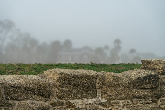 A weathered stone wall stands before a historic building in foggy St. Augustine, Florida. Palm trees and architecture fade into the dense coastal mist.