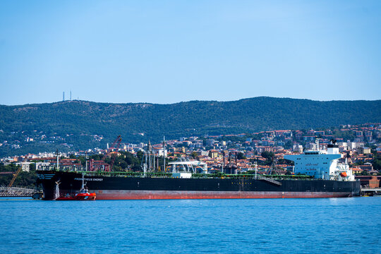 Muggia, Italy - August 8, 2026: Large crude oil tanker Prometheus Energy moored at the industrial terminal in the Gulf of Trieste with cityscape and hills in the background.