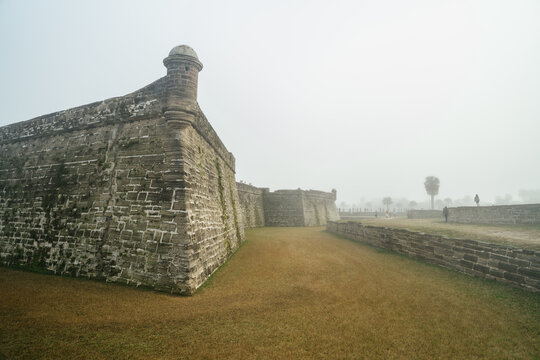 Massive stone walls and a corner watchtower dominate Castillo de San Marcos in St. Augustine, Florida. Tourists walk through the fog around the historic grassy fortress.