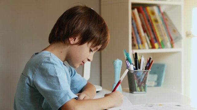 4K slow motion 60fps footage of a concentrated caucasian boy sitting at a white table and writing or drawing with a red marker indoors during daytime.
