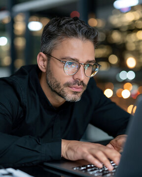 Focused man with glasses types on laptop. Night city lights glow softly in background. His expression shows deep concentration. Ideal for business, tech, or remote work visuals
