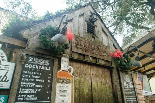 St. Augustine,FL,USA - Dec.30.2024: A rustic wooden St. Augustine Distillery storefront displays a cocktail menu and spirit bottle in Florida. Holiday wreaths, lanterns, and an American flag decorate