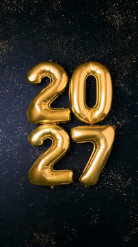 Gold 2027 balloon numbers surrounded by glittering particles on a dark background for New Year celebration. 
