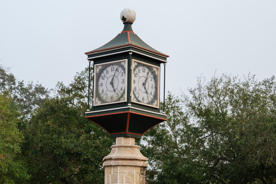 A decorative four-sided clock stands on a stone pedestal in St. Augustine, Florida. The vintage public timepiece rises above green trees against a pale overcast sky.