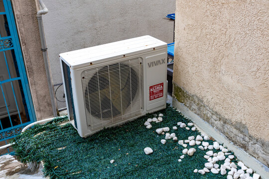 June 30, 2026: Sveti Stefan, Montenegro, White outdoor condenser with visible fan behind metal protective grille mounted on textured stucco wall in narrow urban courtyard, with air, conditioning, cond