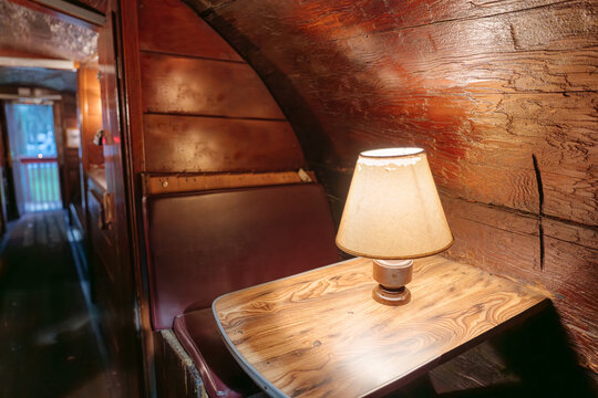 A warm table lamp illuminates a wooden booth with red leather seats inside a vintage passenger train car. The narrow railway compartment features rustic wood walls, a window, and a corridor.
