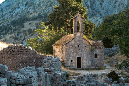 Small stone chapel in mountain ruins with olive trees and dry stone terraces under warm morning sunlight, rocky pathway and weathered masonry in Mediterranean countryside
