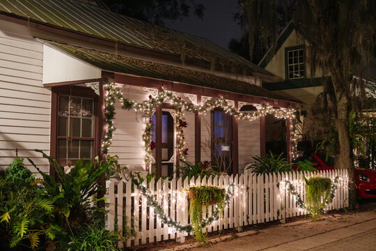 Glowing Christmas lights and garland decorate a small cottage in St. Augustine, Florida. A white picket fence surrounds the tropical garden and illuminated front porch at night.