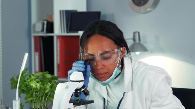 A female scientist wearing a lab coat, safety glasses, gloves, and face mask is closely examining a sample using a microscope in an indoor laboratory setting.