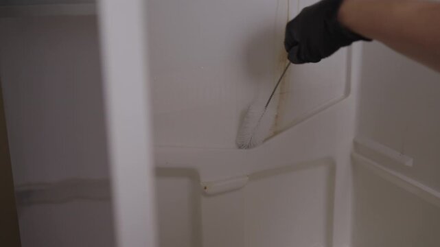 Person in black gloves deep cleaning stubborn brown stain inside refrigerator. Closeup hands scrubbing with brush and wiping surface clean for hygienic home. Concept of household chores and housework.