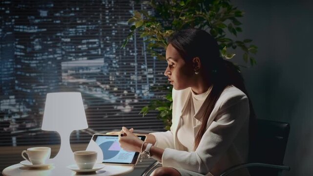 An African American businesswoman in a beige blazer is presenting data on a digital tablet while seated at a table in a modern office with a nighttime cityscape view.