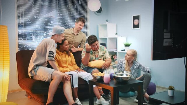 Six adults casually dressed are sitting around a coffee table with snacks in a bright, modern living room featuring a sofa, shelving units, and large windows with blinds.