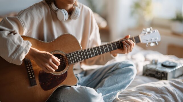 Guitar Serenade at Home: A young person sits on a bed, engrossed in playing an acoustic guitar, with headphones resting around their neck, enjoying a moment of musical bliss in a sunlit room.