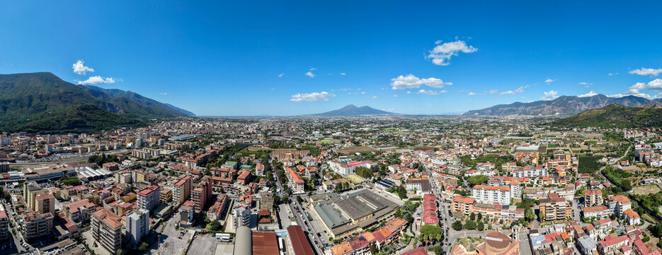 Wide aerial panoramic view of a densely populated Italian town and valley surrounded by green mountains under a blue sky