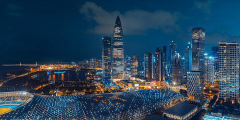 Illuminated Shenzhen skyline with a pointed tower, dense high-rises, waterfront park, bridges, and a curved glass roof beneath clouds at blue hour. © zhao dongfang
