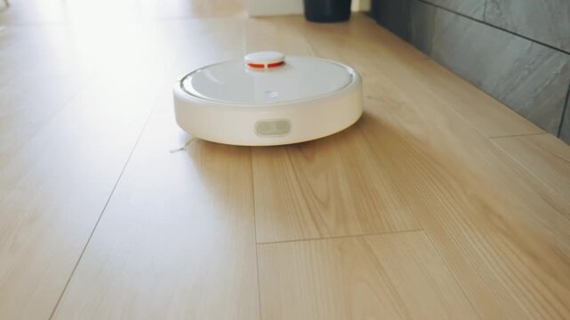close-up white robot vacuum cleaner while cleaning removing dirt dust in home room wooden floor apartment. robotic vacuum moving working at modern interior laminate flooring, parquet, ceramic tiles