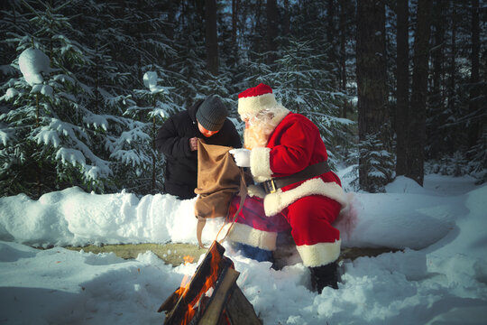 A boy and an authentic Santa Claus with a sack of gifts by a fire in a twilight snowy forest.